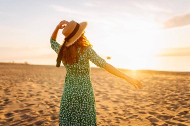 Gün batımı kumsalında saman şapkalı mutlu bir kadının portresi. Elbiseli genç bir kadın deniz manzarasından zevk alıyor, özgürlüğü hissediyor. Etkin yaşam biçimi