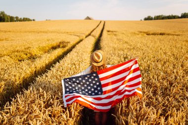 Altın buğday tarlasında şapkalı mutlu kadın Amerikan bayrağı taşıyor. Amerikan bayrağıyla eğlenen ve gün batımında özgürlüğü hisseden genç bir kadın. Kutlama kavramı, özgürlük..