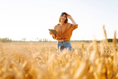 Altın buğday tarlasında çalışan, dijital tabletli gülen kadın tarımcı. Genç kadın çiftçi tarlada mahsul kalitesini kontrol ediyor. Tarım ve teknoloji kavramı.