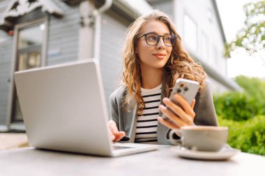 Gözlüklü kadın elinde telefonuyla yazlık bir kafede dizüstü bilgisayarla çalışıyor. Telefon ve dizüstü bilgisayarı olan serbest çalışan kadın güneşli havanın tadını çıkarıyor. Serbest meslek, teknoloji konsepti.