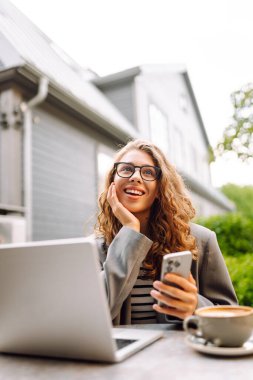 Gözlüklü kadın elinde telefonuyla yazlık bir kafede dizüstü bilgisayarla çalışıyor. Telefon ve dizüstü bilgisayarı olan serbest çalışan kadın güneşli havanın tadını çıkarıyor. Serbest meslek, teknoloji konsepti.