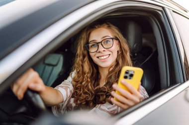 Elinde telefonla araba süren genç bir kadın sürücünün portresi. Telefonu olan güzel bir kadın güneşli bir günde araba sürüyor ve pencereden dışarı bakıyor. Seyahat ve seyrüsefer kavramı.