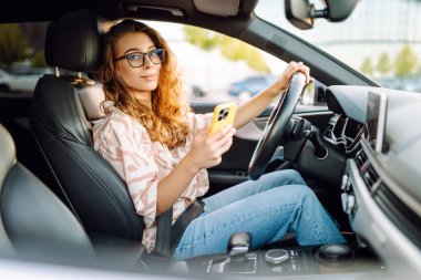 Elinde telefonla araba süren genç bir kadın sürücünün portresi. Telefonu olan güzel bir kadın güneşli bir günde araba sürüyor ve pencereden dışarı bakıyor. Seyahat ve seyrüsefer kavramı.