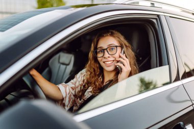Elinde telefonla araba süren genç bir kadın sürücünün portresi. Telefonu olan güzel bir kadın güneşli bir günde araba sürüyor ve pencereden dışarı bakıyor. Seyahat ve seyrüsefer kavramı.