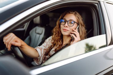 Elinde telefonla araba süren genç bir kadın sürücünün portresi. Telefonu olan güzel bir kadın güneşli bir günde araba sürüyor ve pencereden dışarı bakıyor. Seyahat ve seyrüsefer kavramı.