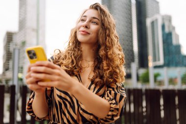 Giyinik ve elinde telefon tutan mutlu kadın şehir caddesinde yürüyor. Sarı telefon blogları ve açık havada mesajlaşan genç bir turist. Turizm, blog konsepti.