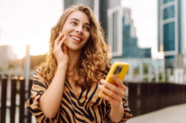 Giyinik ve elinde telefon tutan mutlu kadın şehir caddesinde yürüyor. Sarı telefon blogları ve açık havada mesajlaşan genç bir turist. Turizm, blog konsepti.