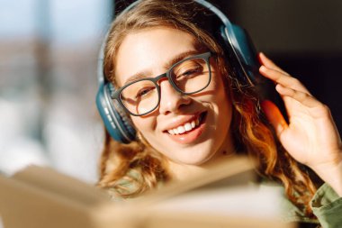 Kablosuz kulaklıklı, rahat bir odada kitap okuyan genç bir kadının yakın çekimi. Müzikten ya da elinde kitapla podcasttan hoşlanan mutlu bir kadın. Rahatlama ve eğitim kavramı.