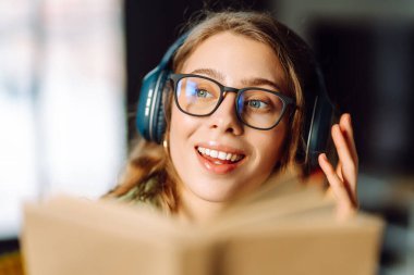 Kablosuz kulaklıklı, rahat bir odada kitap okuyan genç bir kadının yakın çekimi. Müzikten ya da elinde kitapla podcasttan hoşlanan mutlu bir kadın. Rahatlama ve eğitim kavramı.