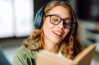 Kablosuz kulaklıklı, rahat bir odada kitap okuyan genç bir kadının yakın çekimi. Müzikten ya da elinde kitapla podcasttan hoşlanan mutlu bir kadın. Rahatlama ve eğitim kavramı.