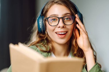 Kablosuz kulaklıklı, rahat bir odada kitap okuyan genç bir kadının yakın çekimi. Müzikten ya da elinde kitapla podcasttan hoşlanan mutlu bir kadın. Rahatlama ve eğitim kavramı.