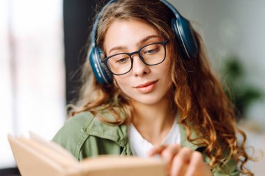 Kablosuz kulaklıklı, rahat bir odada kitap okuyan genç bir kadının yakın çekimi. Müzikten ya da elinde kitapla podcasttan hoşlanan mutlu bir kadın. Rahatlama ve eğitim kavramı.