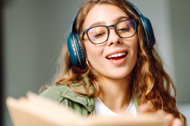 Kablosuz kulaklıklı, rahat bir odada kitap okuyan genç bir kadının yakın çekimi. Müzikten ya da elinde kitapla podcasttan hoşlanan mutlu bir kadın. Rahatlama ve eğitim kavramı.