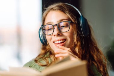 Kablosuz kulaklıklı, rahat bir odada kitap okuyan genç bir kadının yakın çekimi. Müzikten ya da elinde kitapla podcasttan hoşlanan mutlu bir kadın. Rahatlama ve eğitim kavramı.