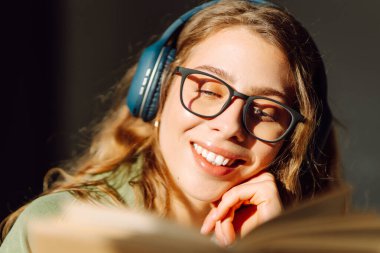 Kablosuz kulaklıklı, rahat bir odada kitap okuyan genç bir kadının yakın çekimi. Müzikten ya da elinde kitapla podcasttan hoşlanan mutlu bir kadın. Rahatlama ve eğitim kavramı.