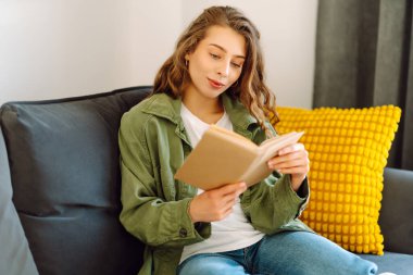 Kitapla gülümseyen kadın rahat bir odada kanepede oturmuş kitap okumaktan zevk alıyor. Genç bir kadın kitap okuyor ve evde dinleniyor. Eğitim, eğlence konsepti. Yaşam biçimi.