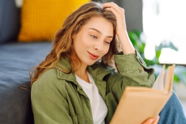 Kitapla gülümseyen kadın rahat bir odada kanepede oturmuş kitap okumaktan zevk alıyor. Genç bir kadın kitap okuyor ve evde dinleniyor. Eğitim, eğlence konsepti. Yaşam biçimi.