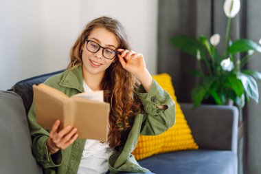 Kitapla gülümseyen kadın rahat bir odada kanepede oturmuş kitap okumaktan zevk alıyor. Genç bir kadın kitap okuyor ve evde dinleniyor. Eğitim, eğlence konsepti. Yaşam biçimi.