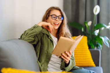 Kitapla gülümseyen kadın rahat bir odada kanepede oturmuş kitap okumaktan zevk alıyor. Genç bir kadın kitap okuyor ve evde dinleniyor. Eğitim, eğlence konsepti. Yaşam biçimi.