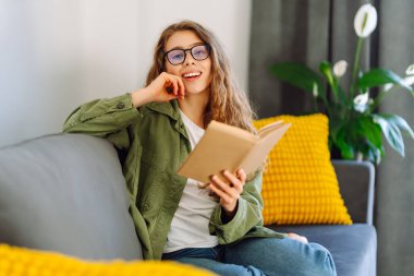 Kitapla gülümseyen kadın rahat bir odada kanepede oturmuş kitap okumaktan zevk alıyor. Genç bir kadın kitap okuyor ve evde dinleniyor. Eğitim, eğlence konsepti. Yaşam biçimi.