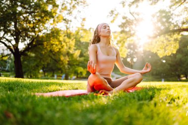 Genç bir kadın parkta minderle yoga yapıyor. Lotus pozisyonunda oturan zinde bir kadın günbatımında huzur ve sükunetin tadını çıkarıyor. Rahatlama kavramı.