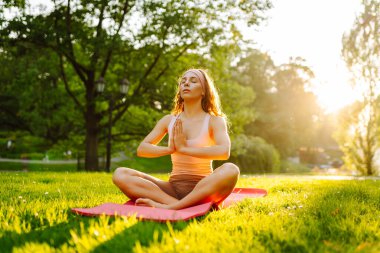 Genç bir kadın parkta minderle yoga yapıyor. Lotus pozisyonunda oturan zinde bir kadın günbatımında huzur ve sükunetin tadını çıkarıyor. Rahatlama kavramı.