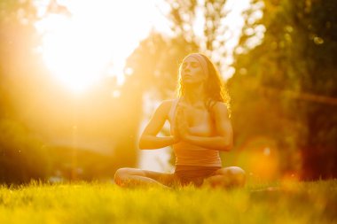 Genç bir kadın parkta minderle yoga yapıyor. Lotus pozisyonunda oturan zinde bir kadın günbatımında huzur ve sükunetin tadını çıkarıyor. Rahatlama kavramı.