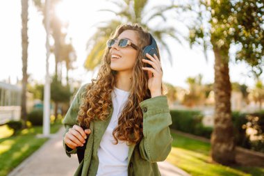 Sırt çantalı ve kablosuz kulaklıklı neşeli kadın güneşli bir parkta müzik dinliyor. Güzel bir turist gün batımını seyrediyor. Yürüyüş, hafta sonu konsepti.