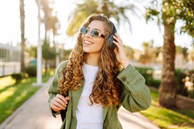Sırt çantalı ve kablosuz kulaklıklı neşeli kadın güneşli bir parkta müzik dinliyor. Güzel bir turist gün batımını seyrediyor. Yürüyüş, hafta sonu konsepti.