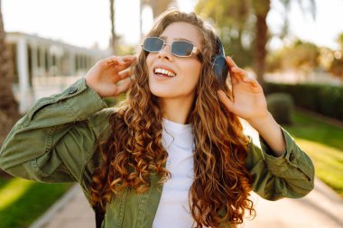 Sırt çantalı ve kablosuz kulaklıklı neşeli kadın güneşli bir parkta müzik dinliyor. Güzel bir turist gün batımını seyrediyor. Yürüyüş, hafta sonu konsepti.