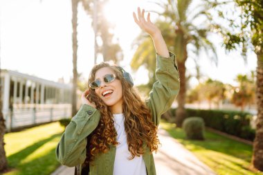 Sırt çantalı ve kablosuz kulaklıklı neşeli kadın güneşli bir parkta müzik dinliyor. Güzel bir turist gün batımını seyrediyor. Yürüyüş, hafta sonu konsepti.