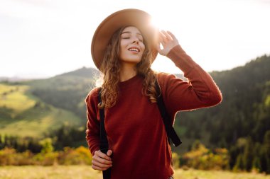 Gün batımında dağ manzarasının tadını çıkaran şapkalı ve sırt çantalı genç bir bayan yürüyüşçü. Güzel bir kadın yemyeşil tepelerde dikilen özgürlüğünü hissediyor. Yürüyüş, macera, özgürlük kavramı.