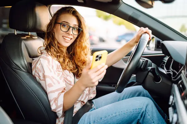 Elinde telefonla araba süren genç bir kadın sürücünün portresi. Telefonu olan güzel bir kadın güneşli bir günde araba sürüyor ve pencereden dışarı bakıyor. Seyahat ve seyrüsefer kavramı.