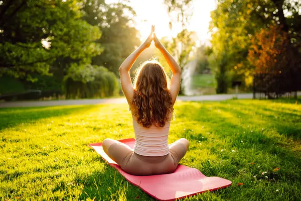 Genç bir kadın parkta minderle yoga yapıyor. Lotus pozisyonunda oturan zinde bir kadın günbatımında huzur ve sükunetin tadını çıkarıyor. Rahatlama kavramı.