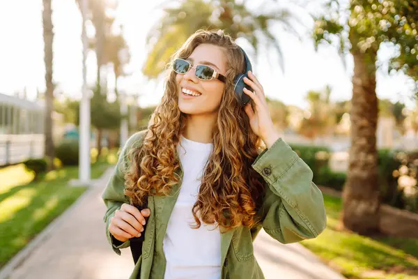 Sırt çantalı ve kablosuz kulaklıklı neşeli kadın güneşli bir parkta müzik dinliyor. Güzel bir turist gün batımını seyrediyor. Yürüyüş, hafta sonu konsepti.