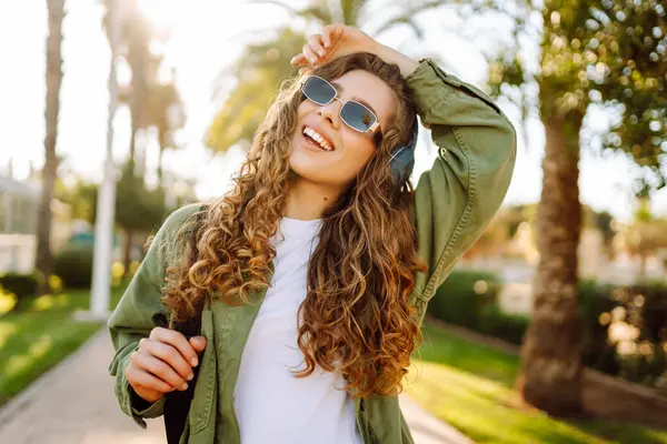 Sırt çantalı ve kablosuz kulaklıklı neşeli kadın güneşli bir parkta müzik dinliyor. Güzel bir turist gün batımını seyrediyor. Yürüyüş, hafta sonu konsepti.