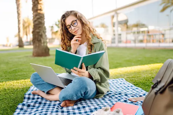 Güneşli bir parkta dizüstü bilgisayarı olan gözlüklü güzel bir kadın. Serbest çalışan bir kadın, dışarıda dizüstü bilgisayarla çalışıyor. Eğitim, uzak çalışma kavramı.