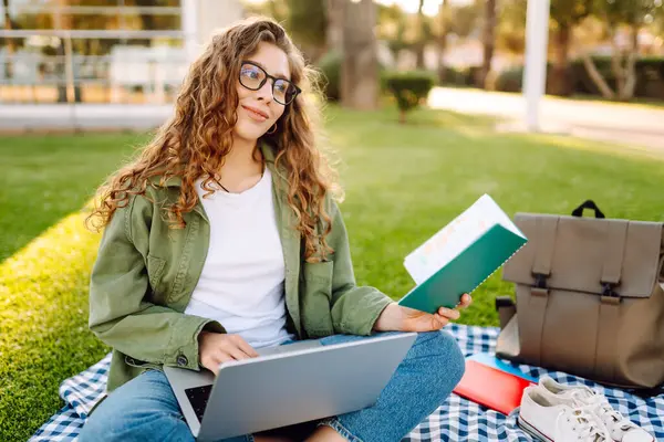 Güneşli bir parkta dizüstü bilgisayarı olan gözlüklü güzel bir kadın. Serbest çalışan bir kadın, dışarıda dizüstü bilgisayarla çalışıyor. Eğitim, uzak çalışma kavramı.