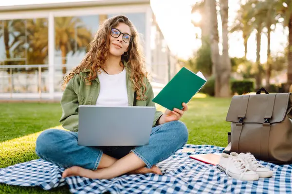 Güneşli bir parkta dizüstü bilgisayarı olan gözlüklü güzel bir kadın. Serbest çalışan bir kadın, dışarıda dizüstü bilgisayarla çalışıyor. Eğitim, uzak çalışma kavramı.