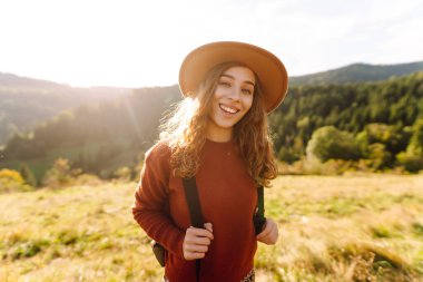Gün batımında dağ manzarasının tadını çıkaran şapkalı ve sırt çantalı genç bir bayan yürüyüşçü. Güzel bir kadın yemyeşil tepelerde dikilen özgürlüğünü hissediyor. Yürüyüş, macera, özgürlük kavramı.