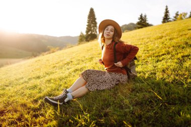 Gün batımında dağ manzarasının tadını çıkaran şapkalı ve sırt çantalı genç bir bayan yürüyüşçü. Güzel bir kadın yemyeşil tepelerde dikilen özgürlüğünü hissediyor. Yürüyüş, macera, özgürlük kavramı.