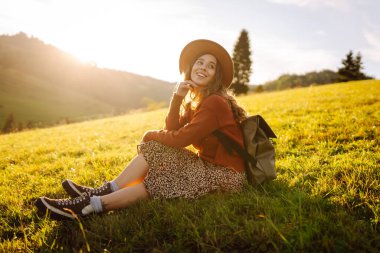 Gün batımında dağ manzarasının tadını çıkaran şapkalı ve sırt çantalı genç bir bayan yürüyüşçü. Güzel bir kadın yemyeşil tepelerde dikilen özgürlüğünü hissediyor. Yürüyüş, macera, özgürlük kavramı.