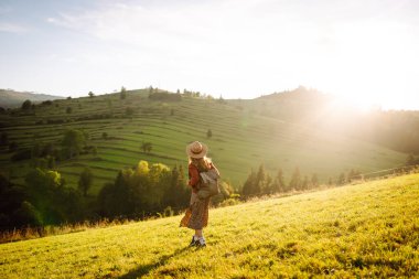Gün batımında dağ manzarasının tadını çıkaran şapkalı ve sırt çantalı genç bir bayan yürüyüşçü. Güzel bir kadın yemyeşil tepelerde dikilen özgürlüğünü hissediyor. Yürüyüş, macera, özgürlük kavramı.