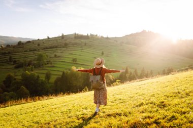 Gün batımında dağ manzarasının tadını çıkaran şapkalı ve sırt çantalı genç bir bayan yürüyüşçü. Güzel bir kadın yemyeşil tepelerde dikilen özgürlüğünü hissediyor. Yürüyüş, macera, özgürlük kavramı.
