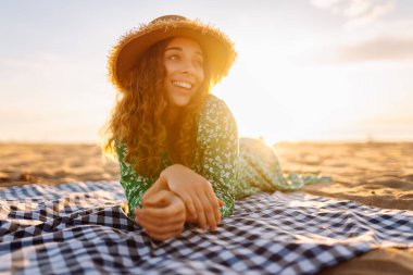 Gün batımında minderde dinlenen şapkalı ve elbiseli güzel bir kadının portresi. Genç bayan deniz kıyısında tatilin tadını çıkarıyor. Özgürlük ve hafta sonu konsepti.