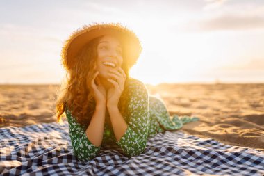 Gün batımında minderde dinlenen şapkalı ve elbiseli güzel bir kadının portresi. Genç bayan deniz kıyısında tatilin tadını çıkarıyor. Özgürlük ve hafta sonu konsepti.