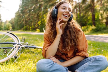 Bisiklet sürdükten sonra çimde oturan kablosuz kulaklıklı genç bir kadın. Güzel bir kadın parkta güneşli bir günün tadını çıkarıyor, müzik dinliyor ve bisiklete biniyor. Etkin yaşam biçimi.