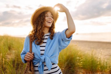Hasır şapkalı neşeli kadın deniz kıyısında dinleniyor. Genç bir kadın sahilde gün batımının tadını çıkarıyor. Özgürlük, rahatlama, hafta sonu konsepti.