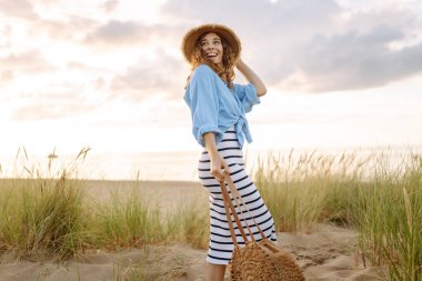 Hasır şapkalı neşeli kadın deniz kıyısında dinleniyor. Genç bir kadın sahilde gün batımının tadını çıkarıyor. Özgürlük, rahatlama, hafta sonu konsepti.