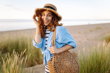 Hasır şapkalı neşeli kadın deniz kıyısında dinleniyor. Genç bir kadın sahilde gün batımının tadını çıkarıyor. Özgürlük, rahatlama, hafta sonu konsepti.